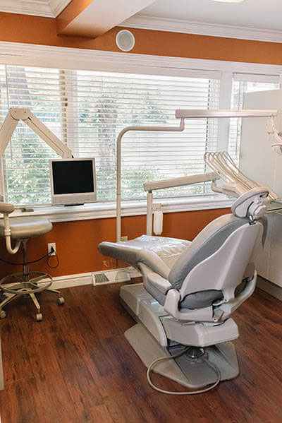 The image shows a dental office interior with a modern dental chair in the foreground, a computer workstation on the left, and a large window allowing natural light into the room.