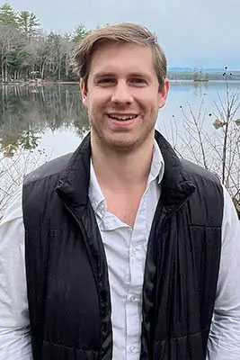 A man standing outdoors with a body of water behind him, smiling at the camera.