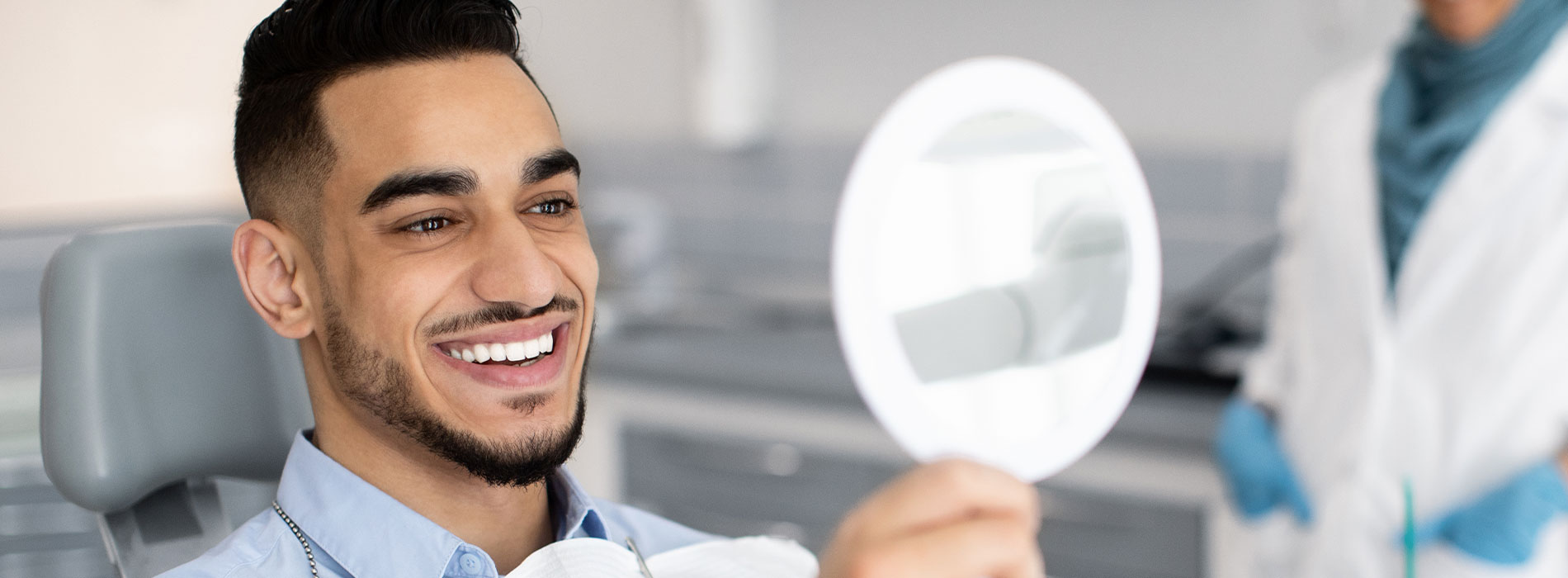 The image shows a smiling man sitting in a dental chair, looking towards the camera with a toothpaste tube held up in front of him.