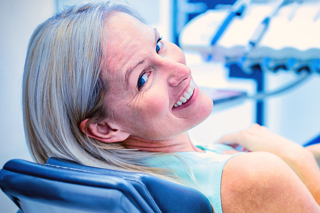 A woman with blonde hair is seated in a dental chair, smiling at the camera.
