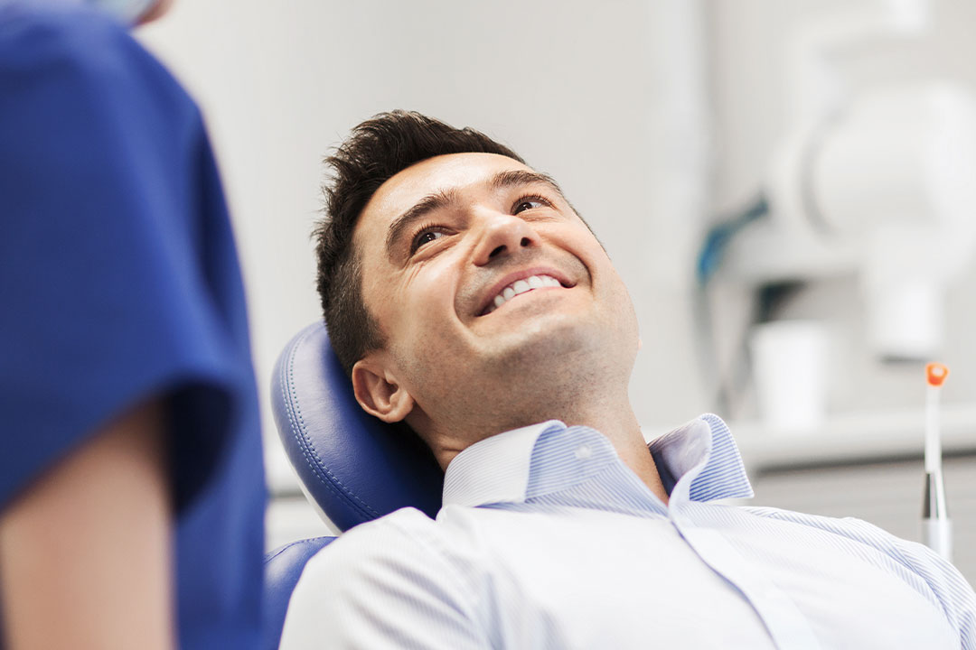 The image shows a man sitting in a dental chair with a big smile on his face, surrounded by dental professionals who appear to be engaged in a positive interaction with him.