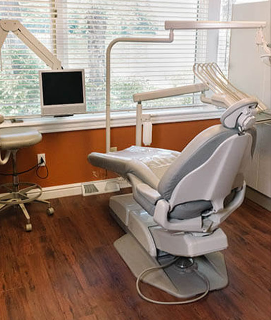 The image shows a dental office interior with a modern dental chair setup, including a dental chair, overhead lighting, and a computer screen displaying patient information, all set against a window with blinds partially drawn.