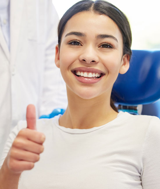 The image shows a young woman with a bright smile giving a thumbs-up gesture while sitting in a dental chair, indicating her satisfaction with a dental visit.