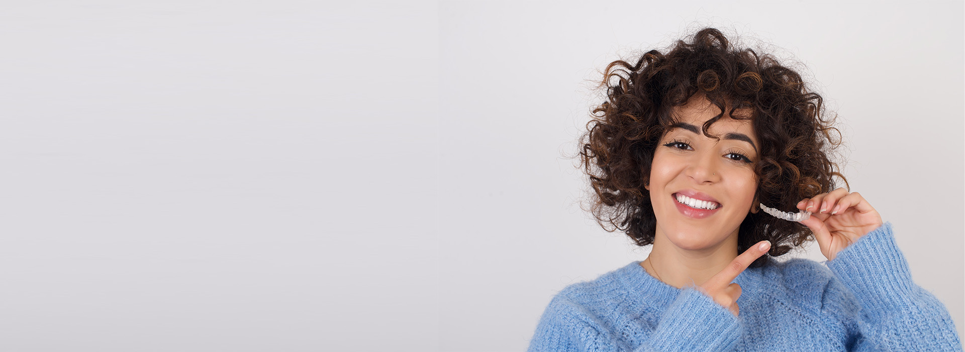 The image features a woman with curly hair holding a toothbrush, smiling at the camera, against a white background.
