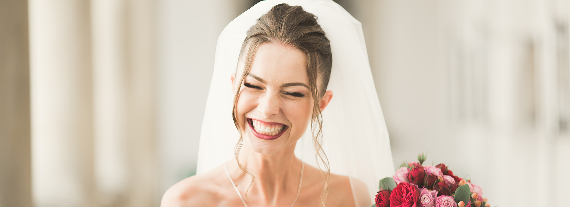 A bride wearing a white veil and holding flowers, smiling at the camera, with her head slightly tilted.
