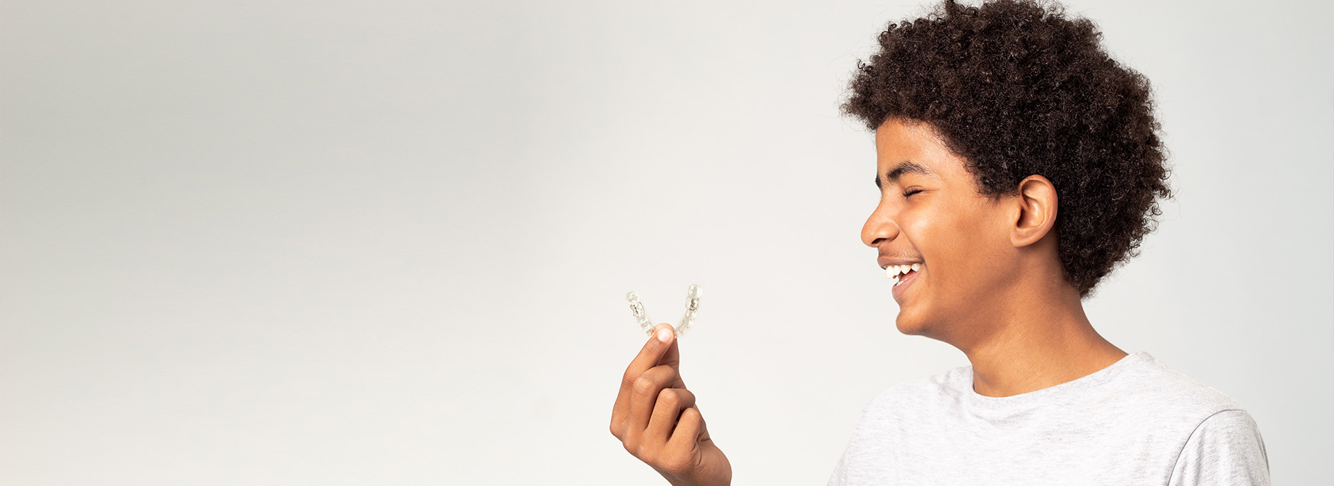 The image shows a young person with curly hair, smiling and holding a small object up to their face, set against a plain background.