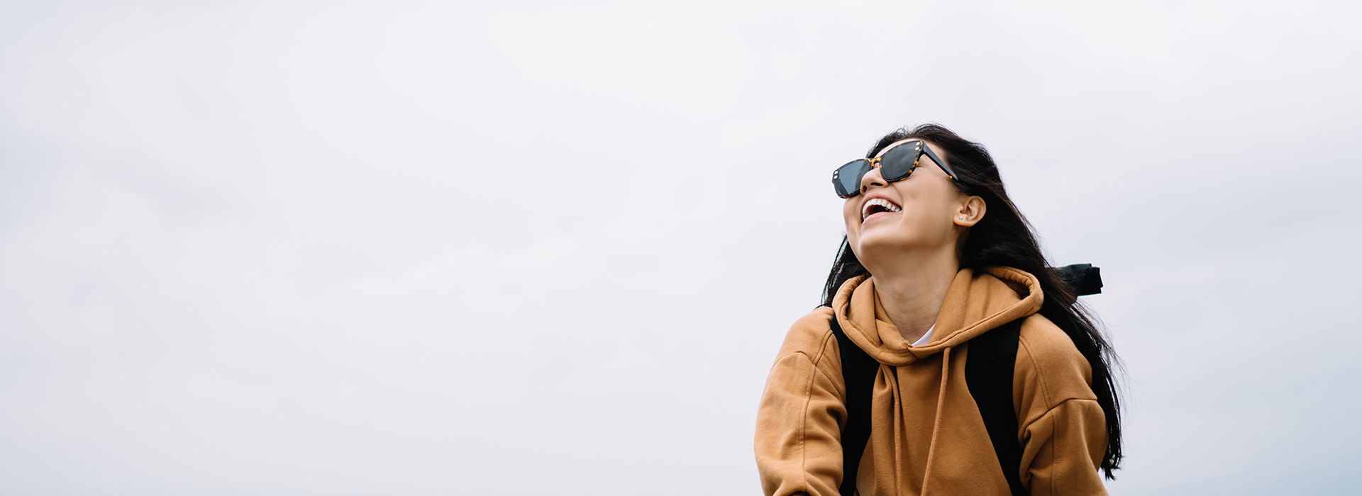 The image shows a person wearing sunglasses, a backpack, and a brown jacket, looking out over a landscape with a clear sky.