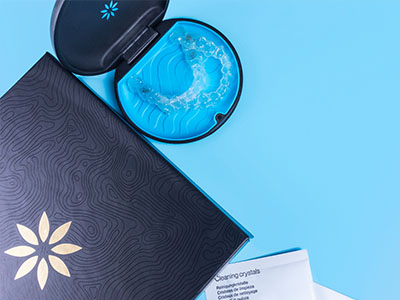 The image displays a blue box with an intricate design on a surface, accompanied by a clear container holding a substance, possibly a product like a candle or incense, with a lid that features a decorative pattern.