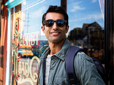 The photo shows a man standing outside a storefront, wearing sunglasses and a backpack, with a smile on his face.