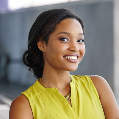 A smiling woman with short hair, wearing a yellow top and standing against a gray background.