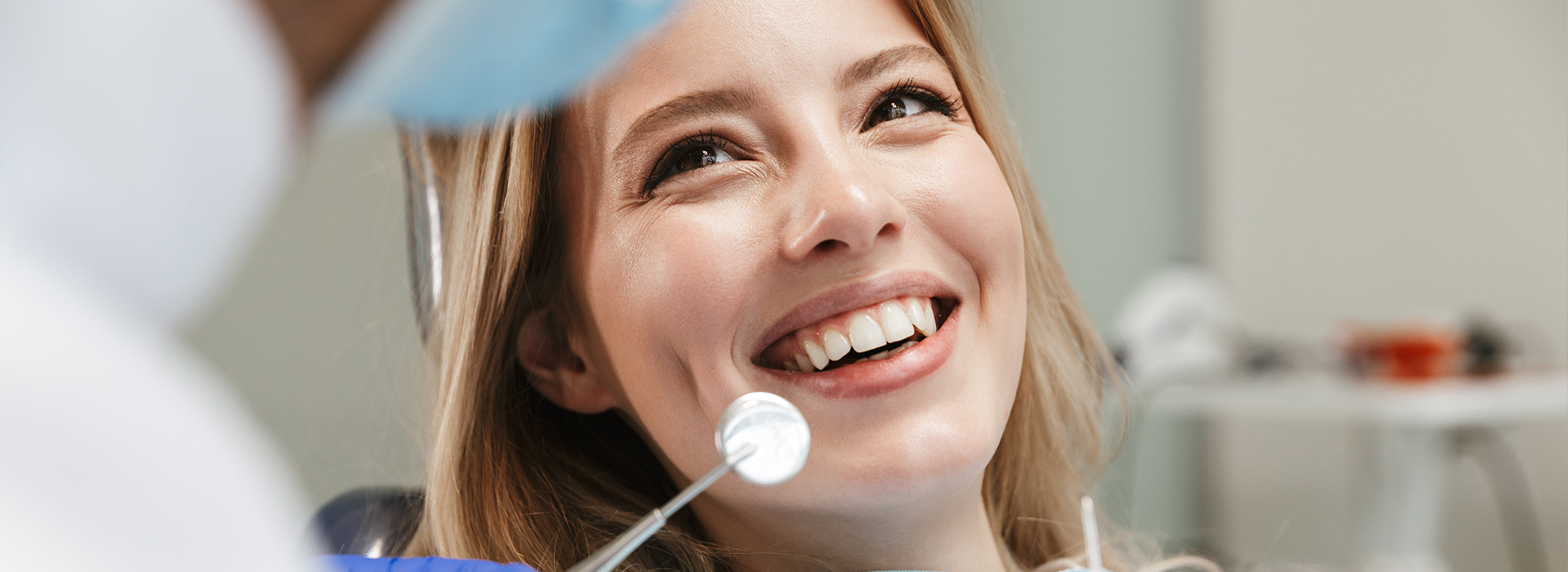 The image shows a woman with blonde hair smiling at the camera while seated in a dentist s chair, with dental tools visible around her, and a person wearing a white coat and stethoscope appears to be examining her.