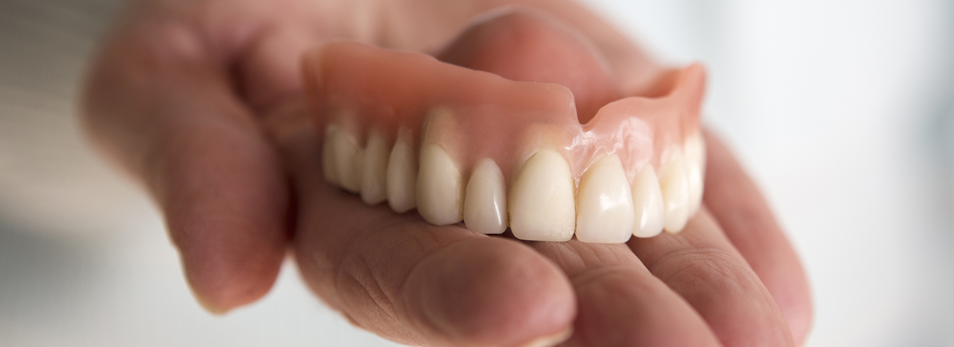 The image shows a human hand holding a set of dentures with visible teeth, displayed against a blurred background.