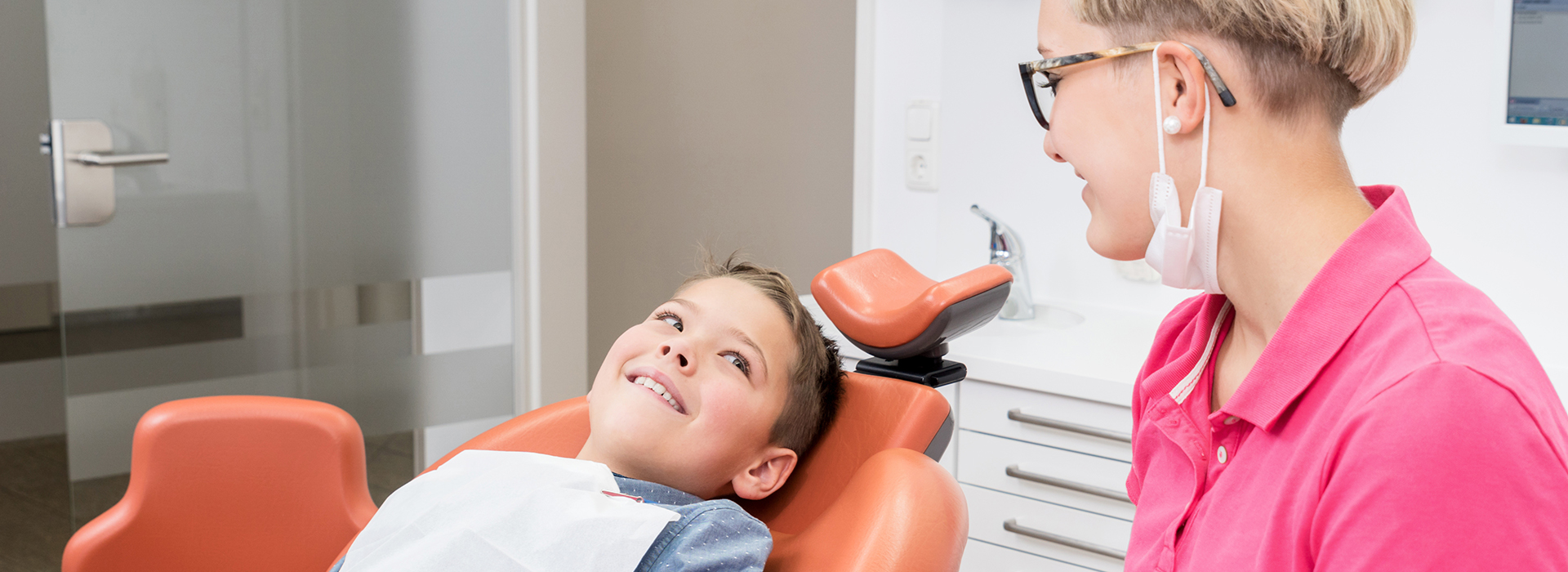 The image shows a young boy sitting in a dental chair with a dentist standing next to him, both smiling at the camera.