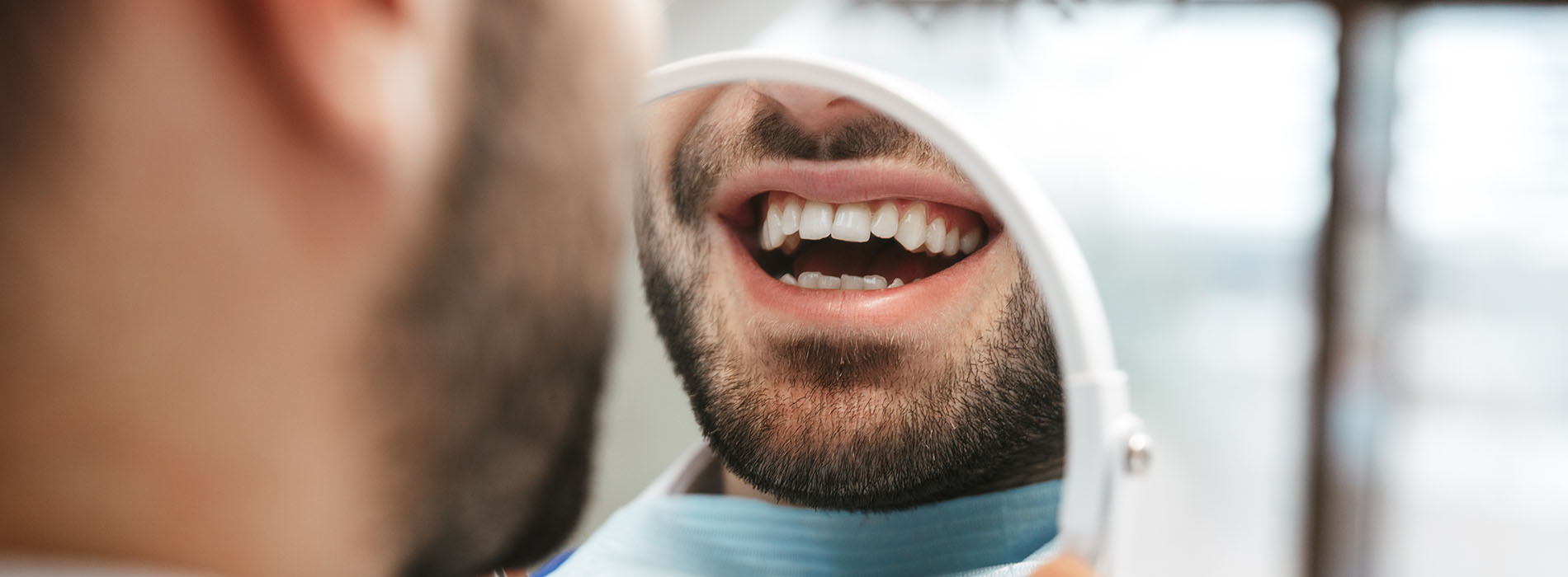 An image featuring a man with a beard seated at a dental chair, looking at his teeth, with a background showing a blurred dentist s office.