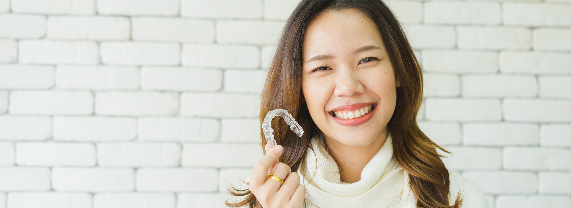 A woman with a bright smile holds up a toothbrush against her teeth, standing in front of a brick wall with a white background.