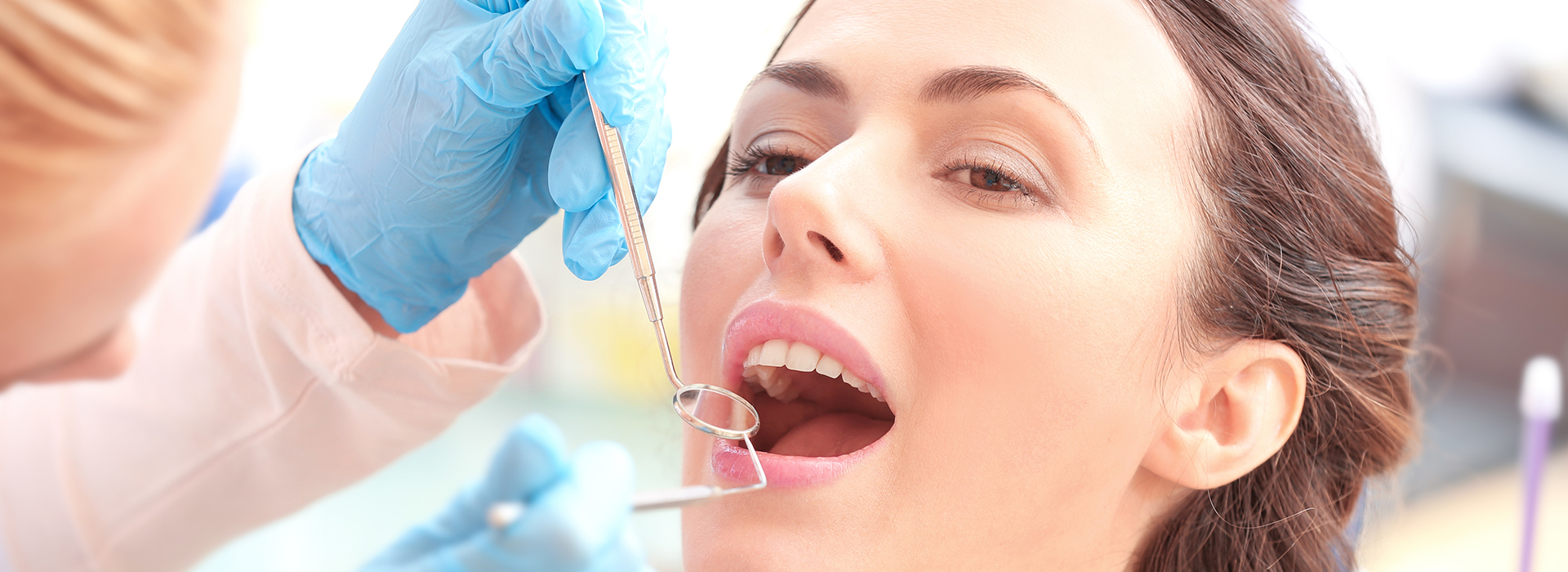 The image shows a woman receiving dental care with a dentist performing a procedure on her teeth while she is seated in a dental chair, surrounded by other dental professionals wearing gloves and masks.