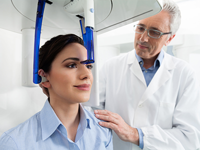 A medical professional stands by a woman seated in a dental chair with a device attached to her head, possibly for dental treatment or examination.