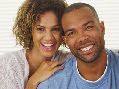 A man and woman stand close together, smiling at the camera, with the man wearing a blue t-shirt and the woman in a white top.