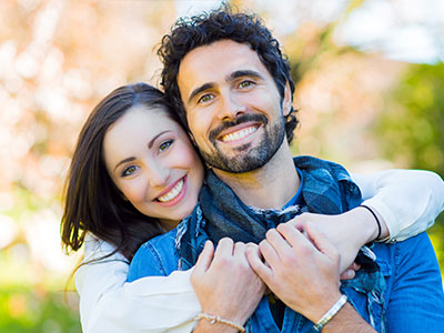 The image shows a man and woman embracing each other with smiles, set against a backdrop of a park-like setting with trees and a clear sky.