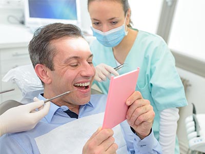 The image shows a man sitting in a dental chair with a pink cardboard sign held up in front of him, smiling while looking at the camera. Behind him, a dental professional is holding a mirror, examining his teeth.