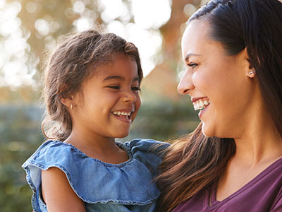 The image depicts a woman and a young child, both smiling, with the woman holding the child. They appear to be outdoors during daylight.