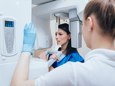 A woman stands next to an advanced medical imaging machine, wearing a white coat and blue gloves, while another person looks on with interest.