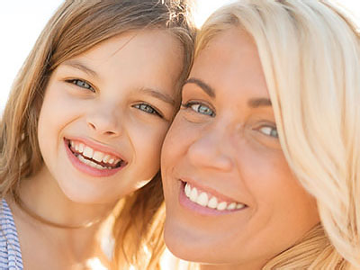 The image shows a woman with a young girl, both smiling and looking at the camera, against a blurred background that suggests an outdoor setting during daylight.