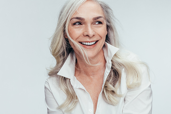 The image features a woman with short hair, wearing a white blouse and smiling at the camera. She has her head slightly tilted to one side and appears to be indoors with a neutral background.