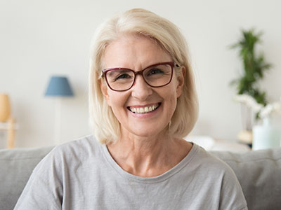 A smiling middle-aged woman with short blonde hair, wearing glasses and a grey top, seated indoors against a white wall.