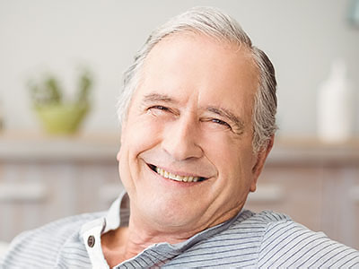 The image shows an older man with grey hair and a smile, wearing a blue shirt, seated comfortably at home.