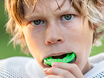 The image shows a young boy with blonde hair, wearing a football uniform, holding a green object in his mouth while looking directly at the camera.