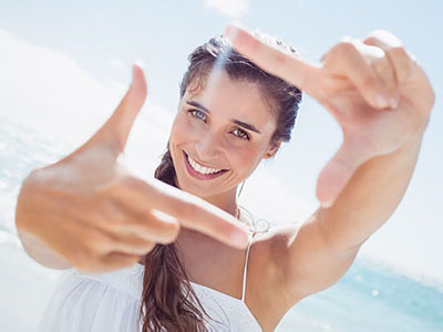 The image features a woman at the beach taking a photo with her hands framing the viewfinder, smiling towards the camera.